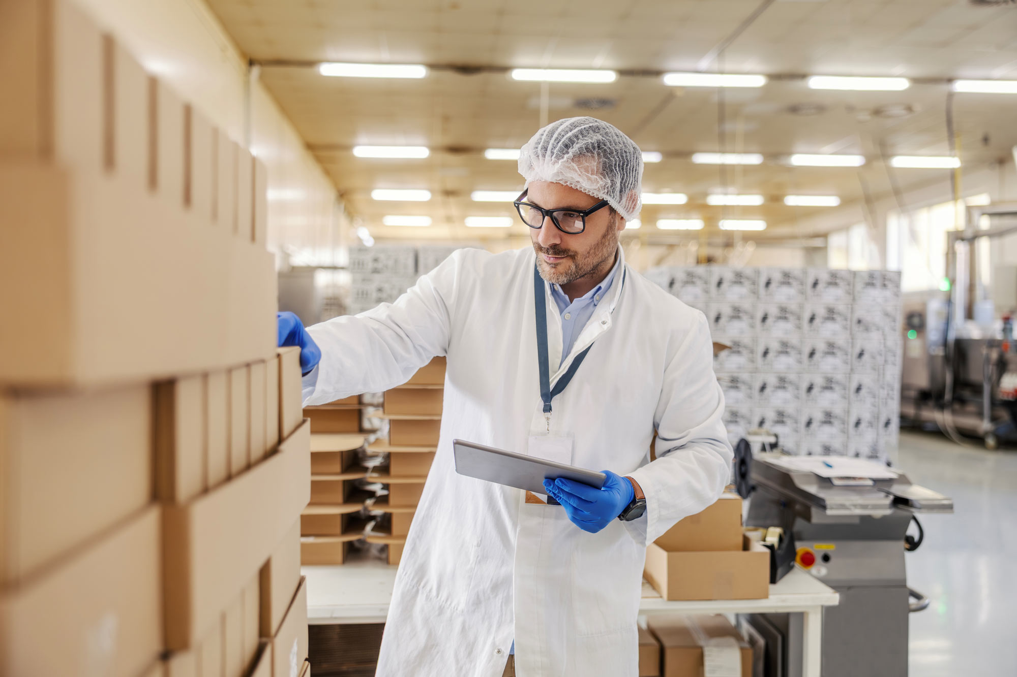 Packaging quality-control specialist inspecting stacked cardboard boxes while recording findings on a tablet in a production facility.
